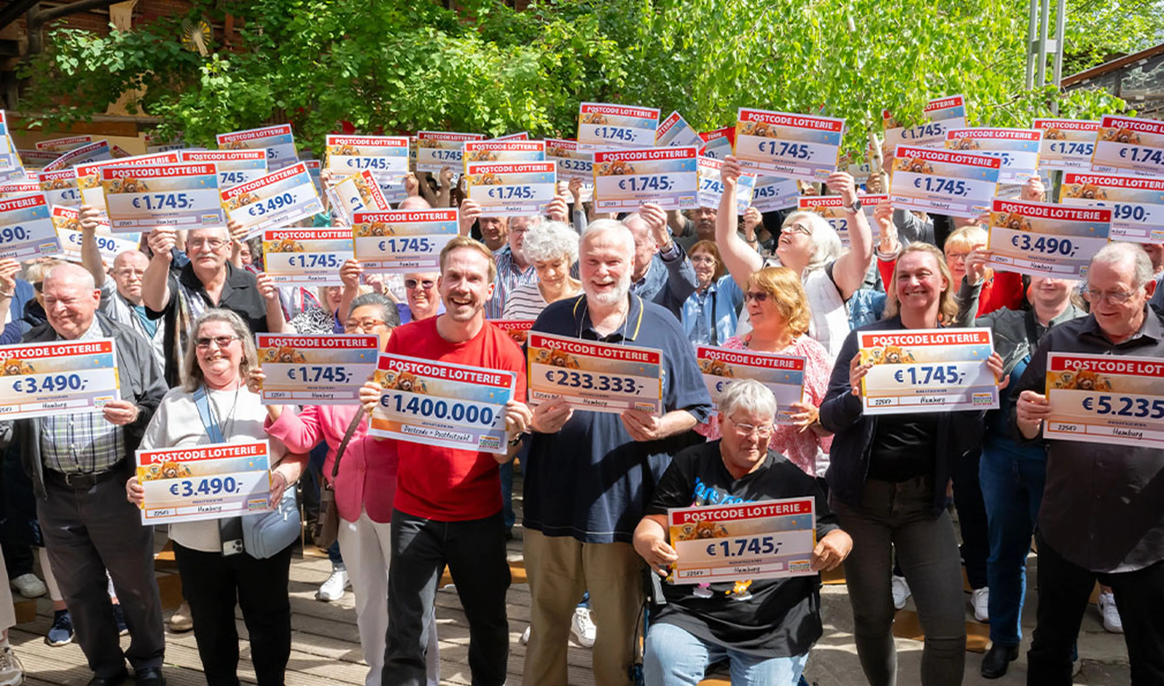 A large group of people holding Postcode Lottery checks outdoors, smiling and celebrating under a sunny, leafy setting.