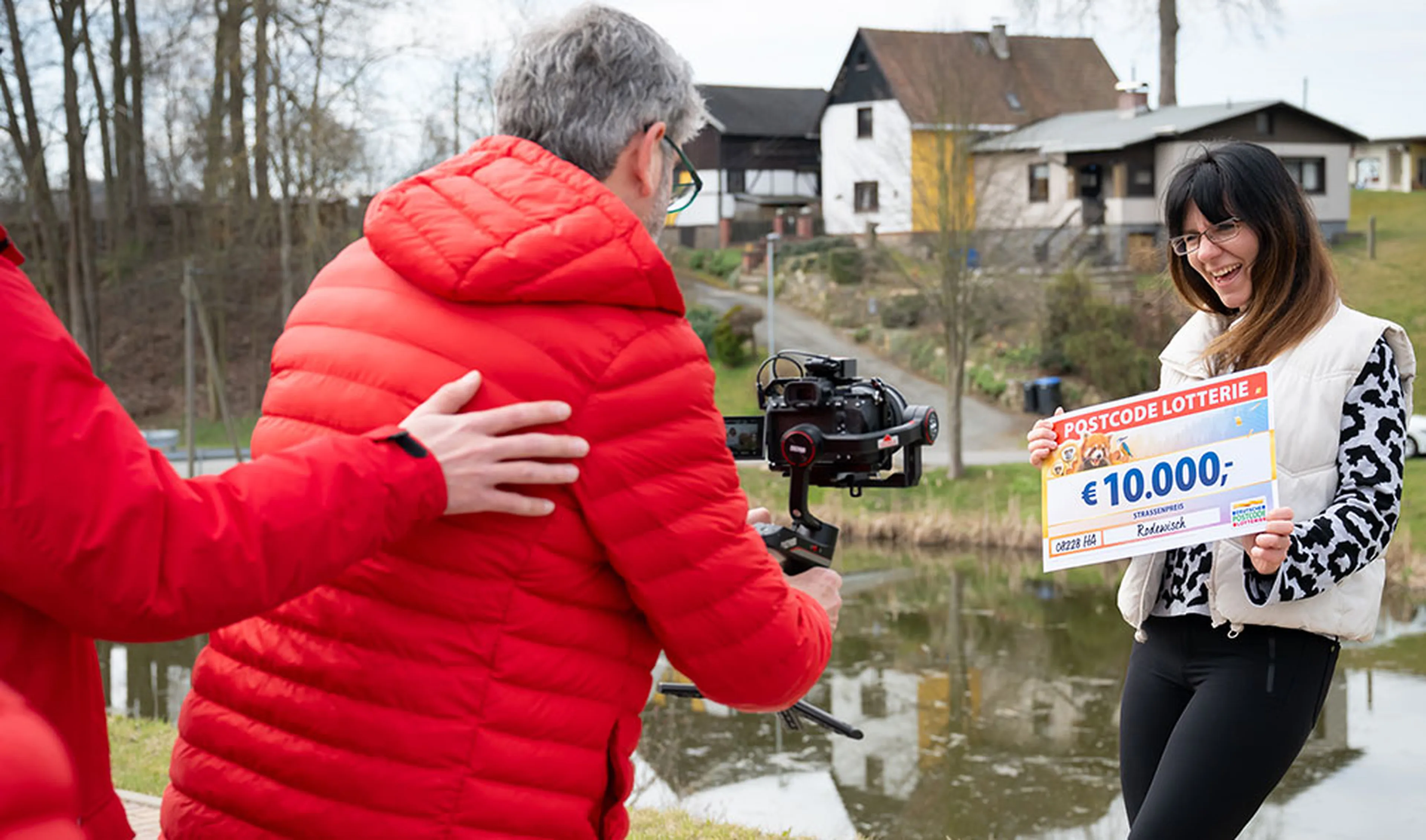 Eine Frau lächelt und hält einen Lotterie-Scheck über 10.000 € in den Händen. Eine Person filmt sie in der Nähe eines Teichs, im Hintergrund sind Häuser zu sehen.