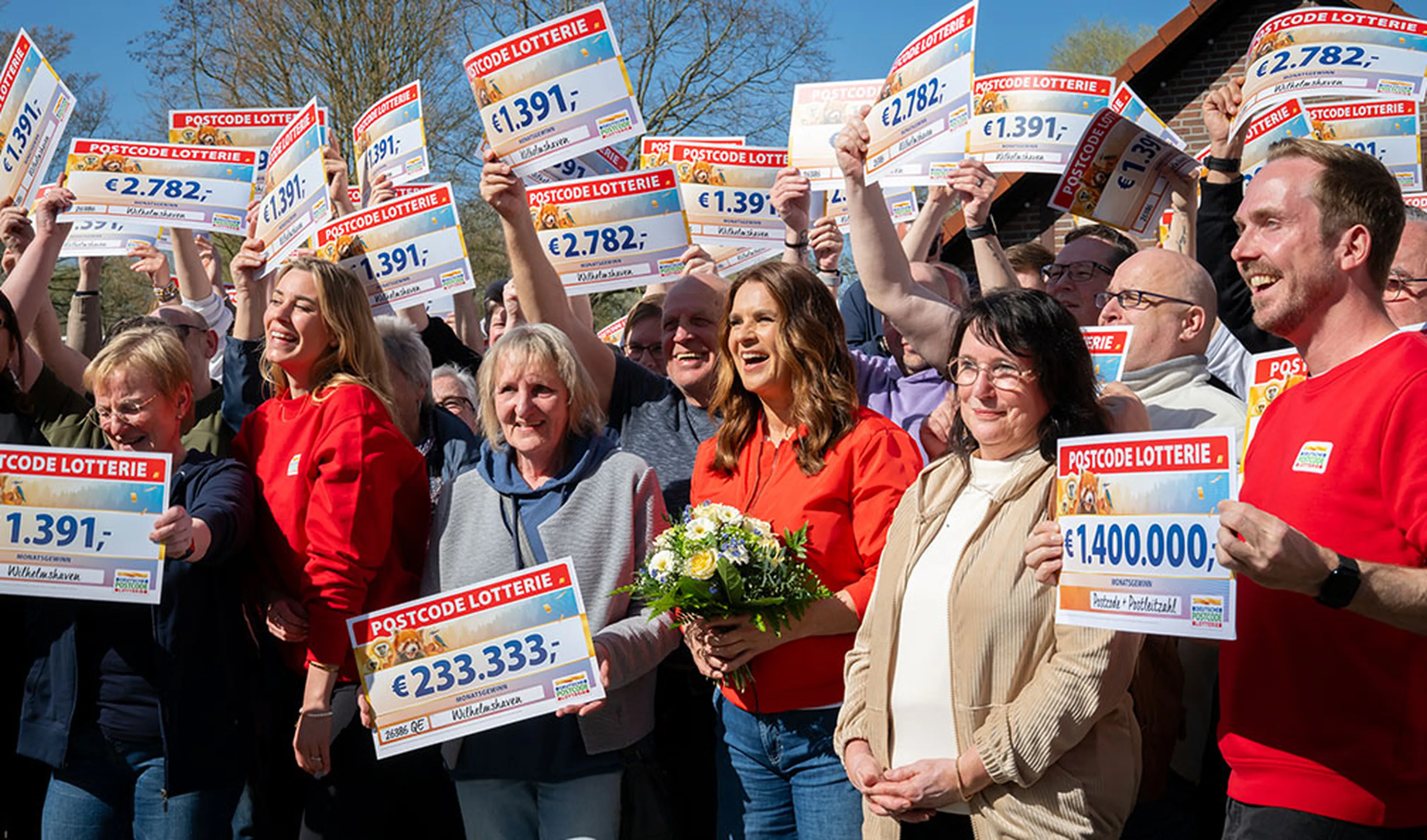 Eine Gruppe von Menschen feiert im Freien und hält große Lotterie-Schecks in den Händen. Eine Frau in Rot trägt lächelnd einen Blumenstrauß und steht inmitten der Menge.
