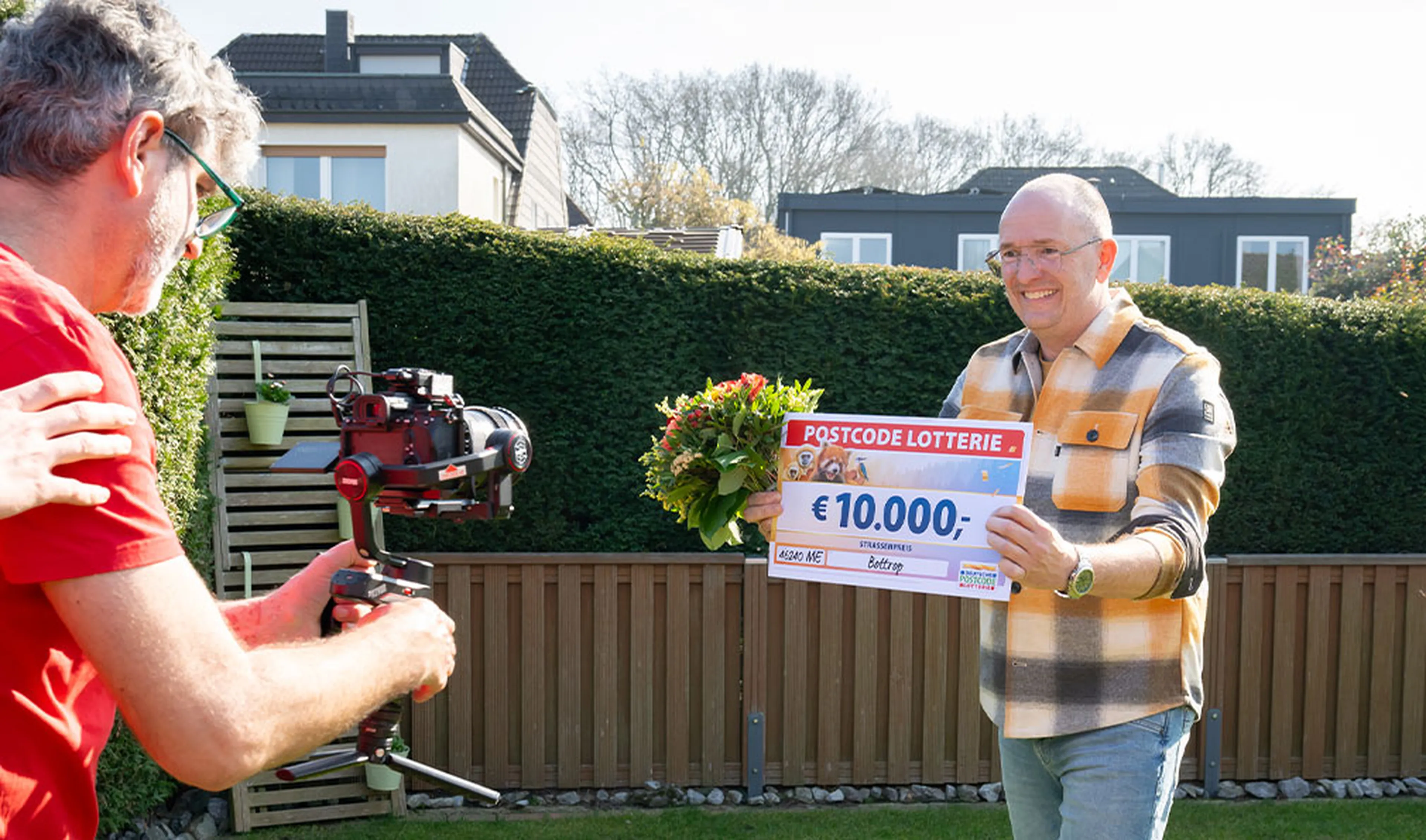 Gewinner zeigt seinen Scheck über 10.000 Euro und hält einen Blumenstrauß in der Hand