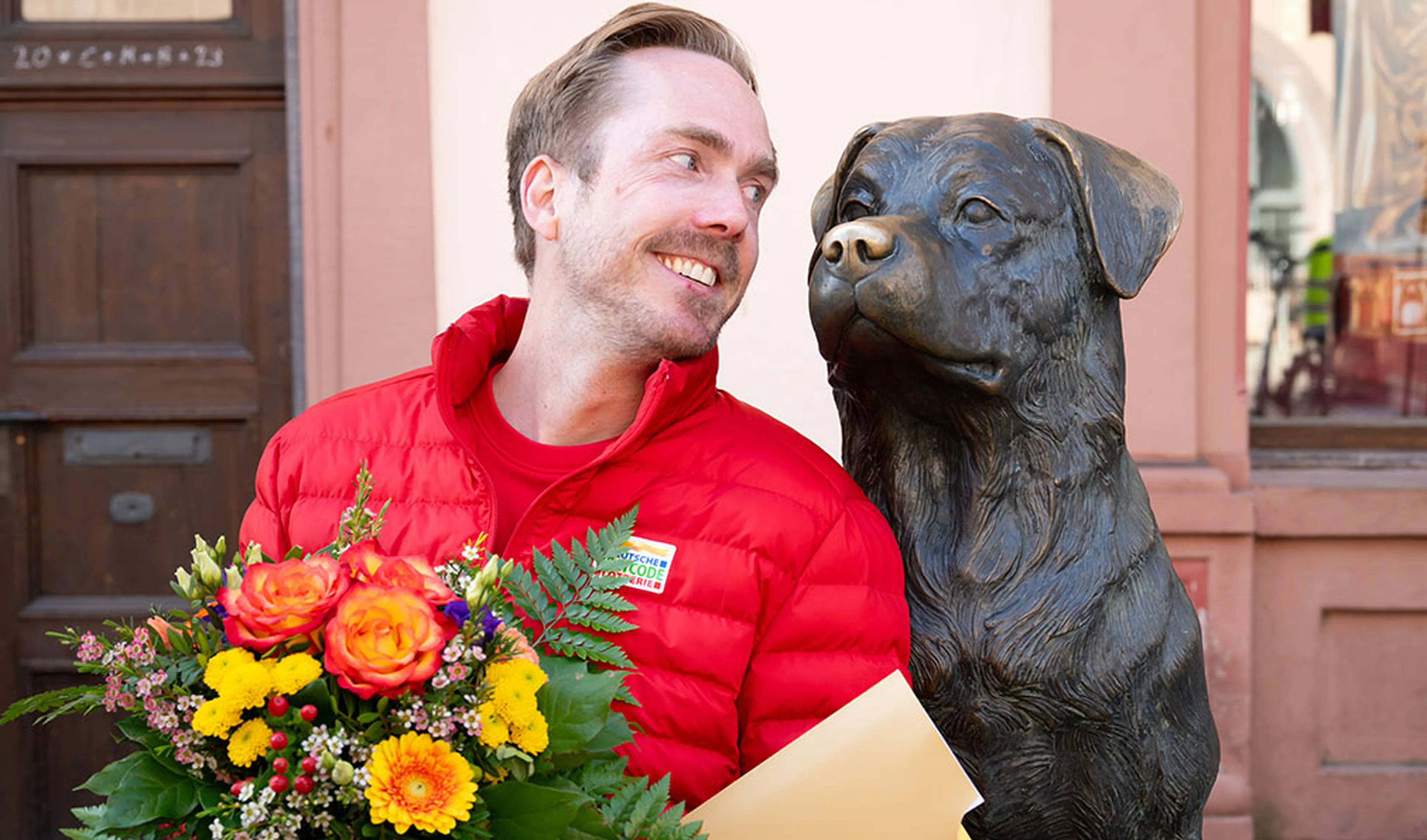 Man in red jacket smiling at a dog statue, holding a bouquet of colorful flowers and an envelope, standing outdoors.