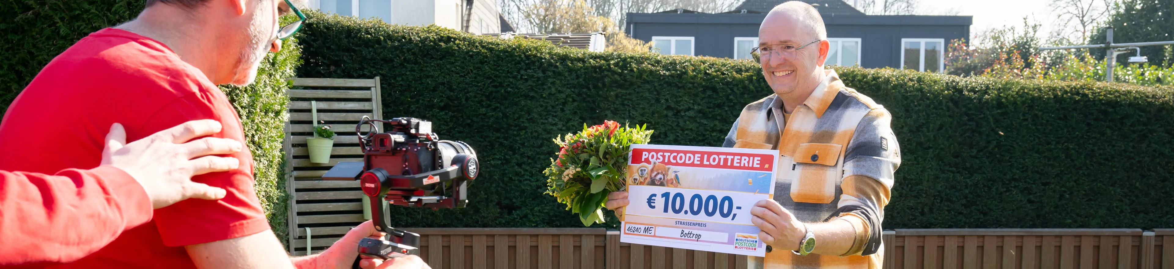 Gewinner zeigt seinen Scheck über 10.000 Euro und hält einen Blumenstrauß in der Hand