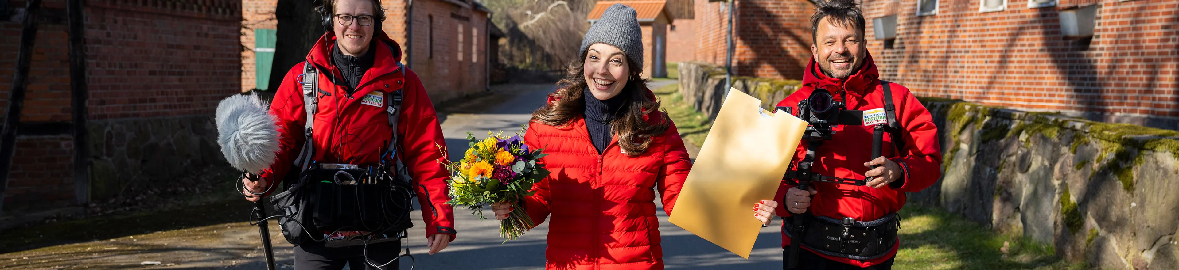Drei Personen in roten Jacken gehen im Freien und lächeln. Eine hält einen Blumenstrauß, eine andere einen großen Umschlag. Entlang des Weges stehen Backsteingebäude.