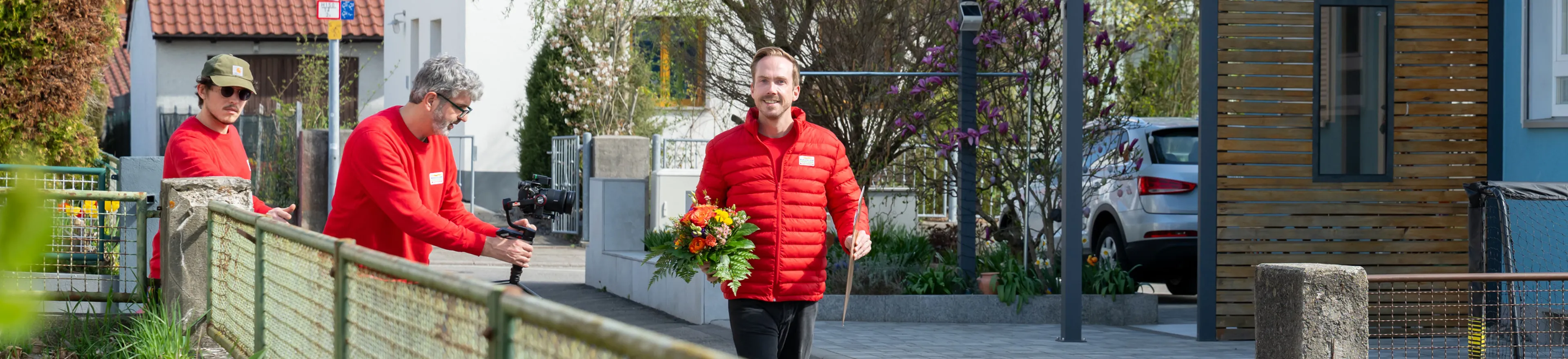 Three people in red jackets are outside. One holds a camera, another carries a bouquet, and the third is walking near a wooden fence.