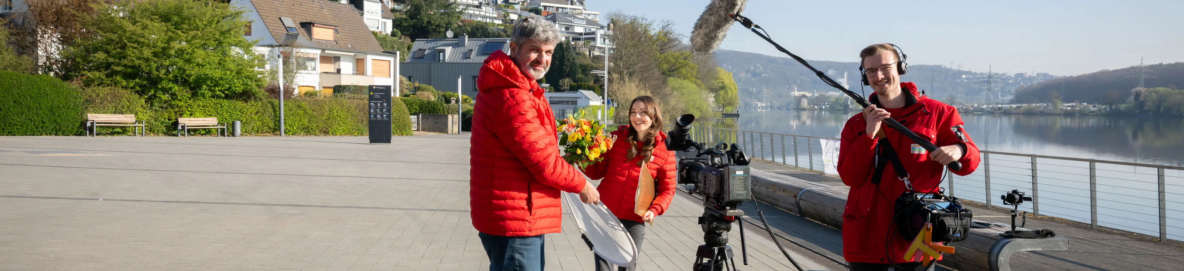 Ein Filmteam in roten Jacken baut Ausrüstung in der Nähe eines Ufers auf. Eine Person hält einen Blumenstrauß in den Händen.