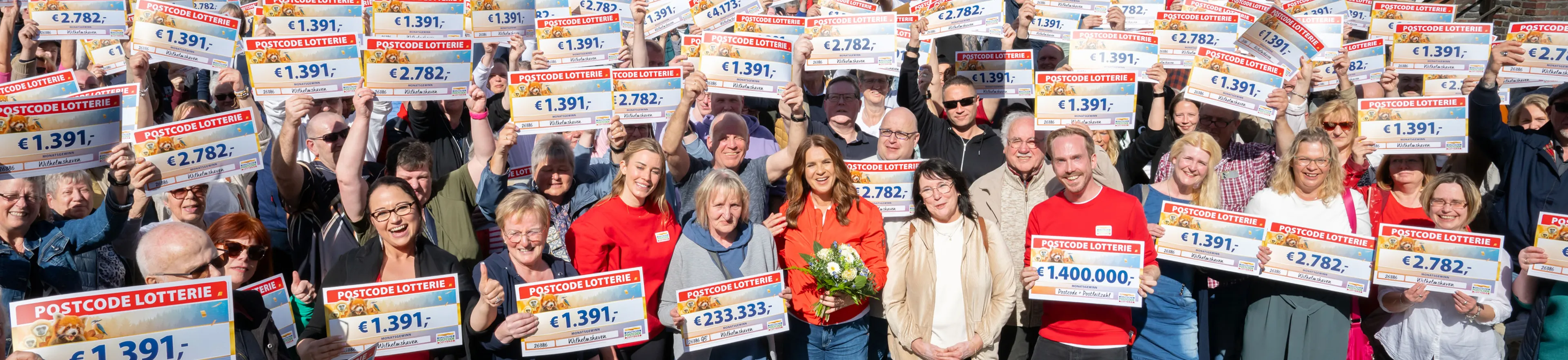 Eine große Gruppe lächelnder Menschen hält im Freien Lotterie-Schecks in den Händen. Einige tragen rote Shirts, und eine Person hält einen Blumenstrauß.