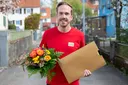 Ein Mann in rotem Shirt hält einen bunten Blumenstrauß und einen großen goldenen Umschlag in den Händen. Er steht im Freien auf einer Wohnstraße.