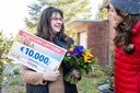 Eine Frau lächelt, hält einen Lotteriescheck über 10.000 € und Blumen in der Hand, während sie sich vor einem Haus mit einer anderen Frau in roter Jacke unterhält.