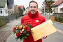 Moderator der Deutschen Postcode Lotterie hält einen goldenen Umschlag und Blumen