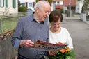 Elderly couple smiling while holding a lottery check and a bouquet on a residential street.