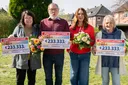 Vier Personen stehen im Freien, lächeln und halten große Lotterie-Schecks über jeweils 233.333 € in den Händen. Sie tragen Blumensträuße und wirken fröhlich.