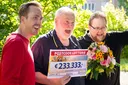 Drei Männer feiern im Freien. Der Mann in der Mitte hält einen Lotterie-Scheck über 233.333 €, der Mann rechts trägt einen bunten Blumenstrauß.