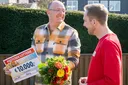Gewinner zeigt seinen Scheck über 10.000 Euro und hält einen Blumenstrauß in der Hand