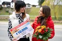 Zwei Frauen lächeln, eine von ihnen hält einen großen Lotterie-Scheck über 10.000 €, die andere einen Blumenstrauß. Im Hintergrund sind Häuser und viel Grün zu sehen.