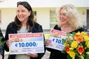 Zwei Frauen lächeln und halten Schecks der Postcode Lotterie über 10.000 € in den Händen. Eine von ihnen trägt einen Blumenstrauß mit gelben und orangefarbenen Blumen.
