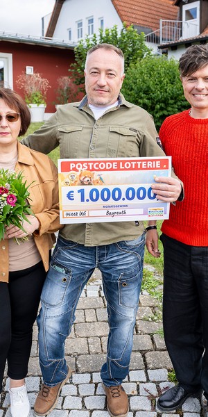Drei Personen stehen im Freien; der Mann in der Mitte hält einen großen Lotterie-Scheck, die Frau links hält Blumen, und der Mann rechts trägt einen roten Pullover.