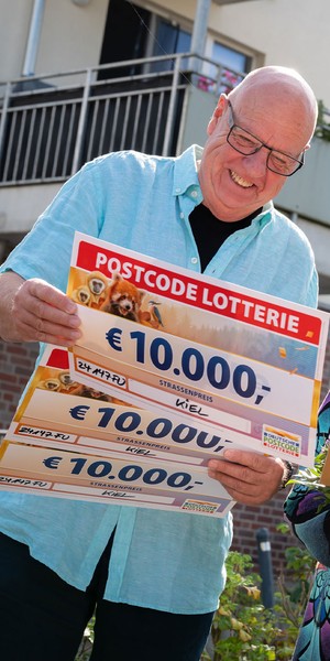 Mann hält im Freien fröhlich große Lotterie-Schecks im Gesamtwert von 10.000 €, neben ihm steht eine Frau mit Blumen.