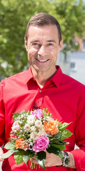 Kai Pflaume lächelt in die Kamera und hat einen Blumenstrauß in der Hand.