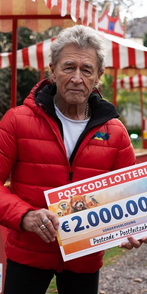 Eine Person in roter Jacke hält ein Schild mit einem Lotteriegewinn über 2.000.000 €. Im Hintergrund ist eine festliche Atmosphäre zu erkennen.