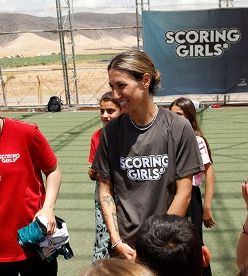 Eine Gruppe junger Mädchen und Frauen versammelt sich auf einem Fußballfeld. Einige tragen Shirts mit der Aufschrift „Scoring Girls“, im Hintergrund ist ein Banner zu sehen.