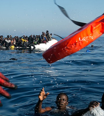 Ein Retter wirft Menschen im Meer eine Schwimmweste zu. Im Hintergrund ist ein überfülltes Boot unter klarem Himmel zu sehen.
