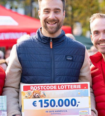 Drei lächelnde Männer in roten Jacken halten einen großen Scheck über 150.000 € von der Postcode Lotterie. Im Hintergrund sind eine Menschenmenge und rote Zelte zu sehen.