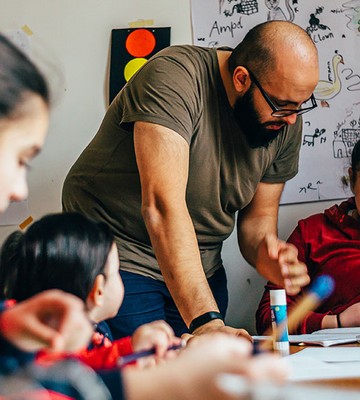 Eine Lehrkraft unterstützt Schülerinnen bei ihrer Arbeit an einem Tisch. Um sie herum liegen Papiere, und an der Wand hängen Kunstwerke. Die Schülerinnen sind konzentriert und engagiert.