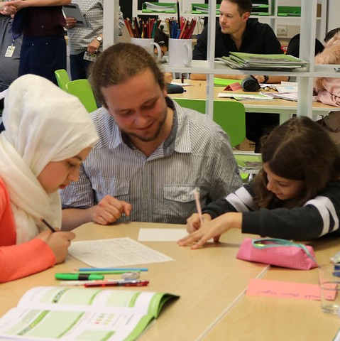 Eine Gruppe von Erwachsenen und Kindern sitzt an einem Tisch und ist in Schreib- und Lernaktivitäten vertieft. Die Szene spielt sich in einem Klassenzimmer ab.