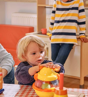 Ein Kind spielt mit einem Spielzeugmixer an einem Tisch, während zwei Erwachsene in einem gemütlichen Raum zuschauen und lächeln.