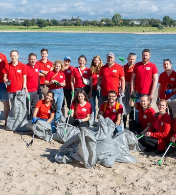 A group of people in red shirts pose with trash bags and pickers on a sandy beach by a river, participating in a cleanup activity.