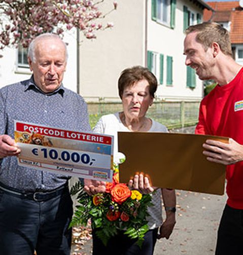 Drei Personen stehen im Freien. Eine von ihnen hält einen großen Scheck über 10.000 € und einen Blumenstrauß, eine andere Person in Rot überreicht einen goldenen Umschlag.
