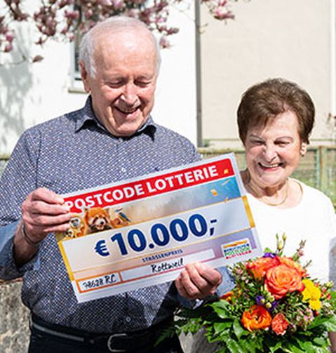 Drei Personen stehen im Freien. Eine von ihnen hält einen großen Scheck über 10.000 € und einen Blumenstrauß, eine andere Person in Rot überreicht einen goldenen Umschlag.