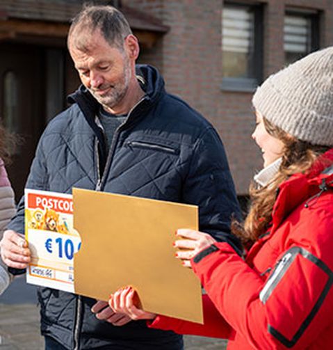 Ein Mann in blauer Jacke hält einen Postcode-Lotterie-Scheck über 1.000 € und steht im Freien neben einer Frau in rotem Mantel und grauer Mütze.