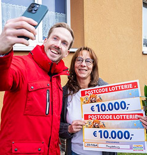 Ein Mann in roter Jacke macht ein Selfie mit einer Frau, die zwei Postcode-Lotterie-Schecks über jeweils 10.000 € hält. Sie stehen vor einem Haus.