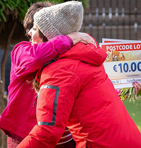 Zwei Personen umarmen sich im Freien. Eine trägt einen roten Mantel, die andere eine rosafarbene Jacke und hält einen großen Lotterie-Scheck über 10.000 € in den Händen.