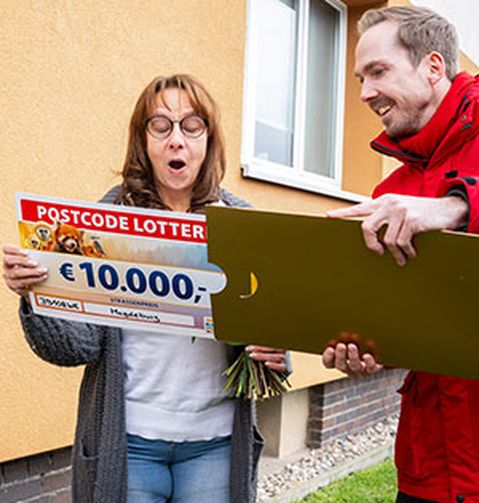 Eine Frau ist überrascht über einen Lotterie-Gewinn von 10.000 € und steht neben einem lächelnden Mann, der eine große goldene Karte hält. Sie befinden sich vor einem Gebäude.