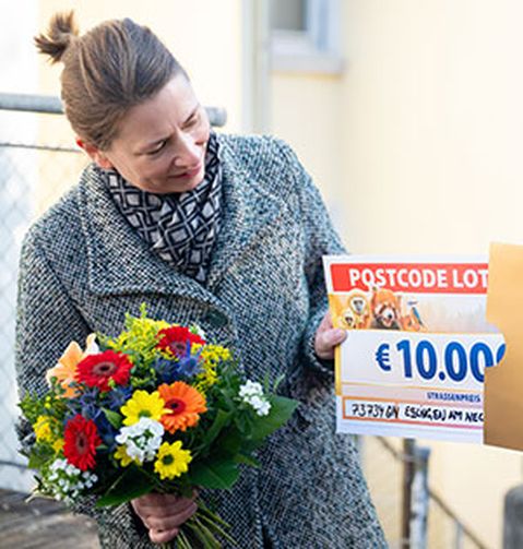 Eine Frau hält einen bunten Blumenstrauß und einen Lotterie-Scheck über 10.000 € in der Hand und lächelt im Freien.