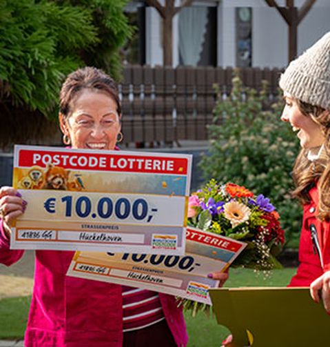 Zwei Frauen lächeln, die eine hält einen großen Scheck über 10.000 € von der Postcode Lotterie, die andere einen Blumenstrauß.