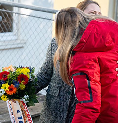 Zwei Personen umarmen sich im Freien. Eine von ihnen hält einen bunten Blumenstrauß und eine große Karte. Im Hintergrund ist ein Maschendrahtzaun zu sehen.