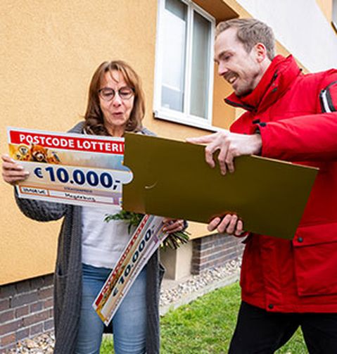 Eine Frau ist überrascht über einen Lotterie-Gewinn von 10.000 € und steht neben einem lächelnden Mann, der eine große goldene Karte hält. Sie befinden sich vor einem Gebäude.