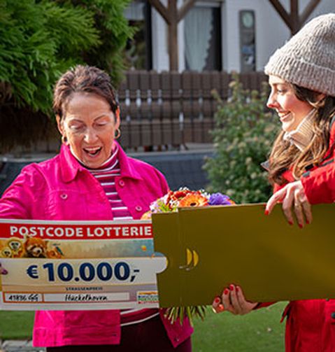 Zwei Frauen lächeln im Freien. Eine von ihnen hält einen großen Lotterie-Scheck über 10.000 € sowie einen Blumenstrauß.