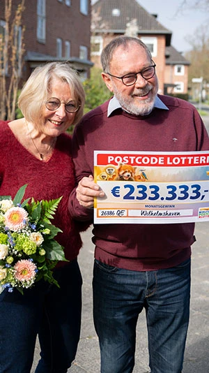Ein älteres Paar lächelt, hält einen Lotterie-Scheck über 233.333 € und einen Blumenstrauß in den Händen. Sie stehen im Freien in einem Wohngebiet.
