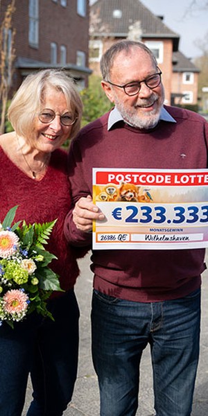 Ein älteres Paar lächelt, hält einen Lotterie-Scheck über 233.333 € und einen Blumenstrauß in den Händen. Sie stehen im Freien in einem Wohngebiet.