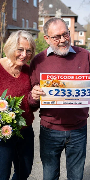 Ein älteres Paar lächelt, hält einen Lotterie-Scheck über 233.333 € und einen Blumenstrauß in den Händen. Sie stehen im Freien in einem Wohngebiet.