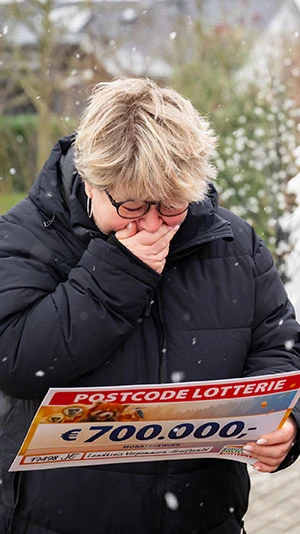 Eine Person in schwarzem Mantel reagiert emotional, während sie einen Postcode-Lotterie-Scheck über 700.000 € in den Händen hält. Die Szene spielt im verschneiten Freien.
