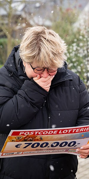 Eine Person in schwarzem Mantel reagiert emotional, während sie einen Postcode-Lotterie-Scheck über 700.000 € in den Händen hält. Die Szene spielt im verschneiten Freien.