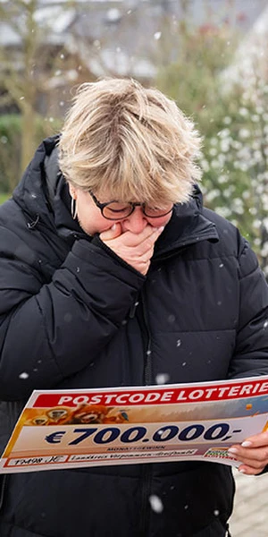 Eine Person in schwarzem Mantel reagiert emotional, während sie einen Postcode-Lotterie-Scheck über 700.000 € in den Händen hält. Die Szene spielt im verschneiten Freien.