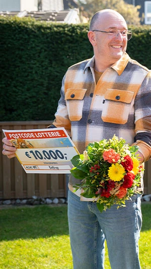 Ein Mann lächelt, hält einen Postcode-Lotterie-Scheck über 10.000 € und einen Blumenstrauß in den Händen. Er steht in einem Garten.