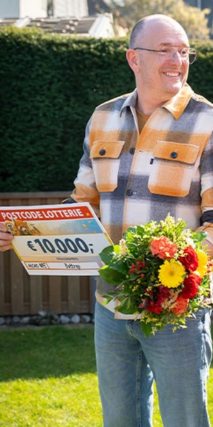Ein Mann lächelt, hält einen Postcode-Lotterie-Scheck über 10.000 € und einen Blumenstrauß in den Händen. Er steht in einem Garten.