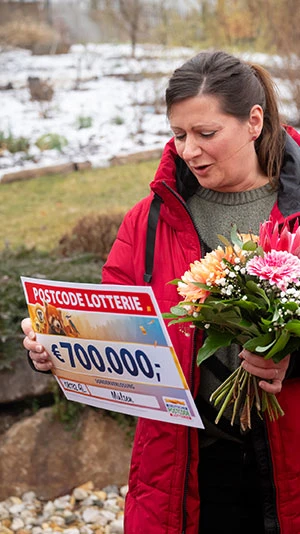 Eine Frau in rotem Mantel hält einen Blumenstrauß und einen großen Scheck über 700.000 € von der Postcode Lotterie. Sie steht im Freien vor einer verschneiten Kulisse.